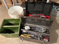 Two black metal tool boxes filled with hand tools, a green plastic container, a white bucket on the floor