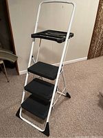 Photo of white metal ladder with black textured steps and built-in tray, standing on carpet.