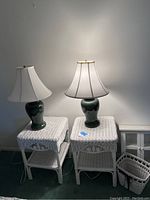 Two white wicker bedside tables each with a green ceramic base lamp and fabric shade on top, side by side on carpeted floor against wall.