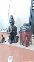 Photo showing all three Buddha statues together on the floor: small pottery Buddha, tall wood kneeling Buddha, and large metal Buddha head.