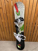 Snowboard upright against wood backdrop with attached bindings showing top sheet
