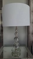 Full view of a glass table lamp showing its white drum shade and stacked spherical glass design.