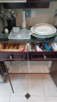 Full view of kitchen drawers with cutlery and assorted utensils, plates stacked on countertop, and utensil holders.