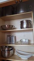 Large stainless steel pots on top shelf and mixing bowls, graters, white serving dishes, and glass baking dishes on lower shelves of wooden cabinet