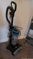 Full view of Bissell upright vacuum and partially visible gray metal step ladder against wall on carpet.