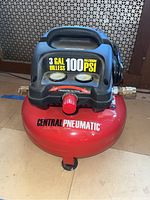 Top and front view of Central Pneumatic oil-less air compressor showing 3 gallon capacity and 100 PSI max pressure with pressure gauges and red control knob.