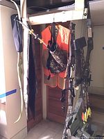 Two bows hanging: green recurve practice bow at left and camouflage compound bow at right, with attached arrow quiver and orange vest in background.