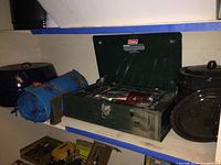Photo showing green Coleman camp stove with two burners, black enamel roasting pan, large black steam pot and blue rolled camping mat on shelf.