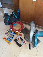 Photo of all items in lot including Makita circular saw, air brad nailer, red air hose, boxes of brads, bottle of tool oil, and carrying bag on floor.