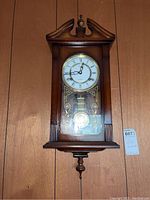 Front view of the wooden wall clock showing the round clock face with Roman numerals and decorative pendulum behind the glass door.