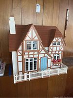Front view of wooden Tudor style dollhouse with white picket fence and detailed windows and door resembling a small cottage.