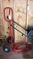 Full view of red hand truck dolly leaning against plywood wall with orange extension cord coiled at base.