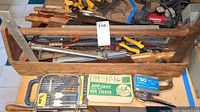 Photo showing wooden-handled hand saws, hacksaws, a gun tacker, drill bits case, and some other hand tools in a wooden toolbox.