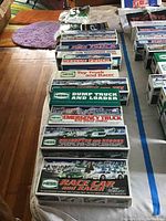 Photo shows eleven Hess trucks in their original boxes arranged on a table, some stacked. Boxes include models like race car, rescue, helicopter rescue, and emergency truck, featuring colorful illustrated winter backgrounds.