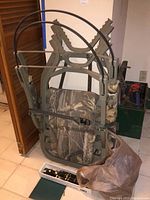 Hunting chair in camouflage attached to a tree with straps with an open plastic bag and knife box on floor nearby.