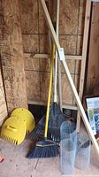 Overall grouping of lawn care tools including rakes, broom, leaf collection paddles, and wire mesh rolls in a storage shed area.