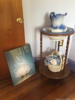 Porcelain wash basin and pitcher set on wooden stand with basket below and wall canvas leaning nearby