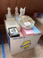 Box of Precious Moments figurines including a child holding a flag, a sitting child, and the decorative plaque on top