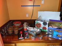 Wide view showing the lot contents on a wooden dresser including dishes, glassware, kitchen tools, and boxed items