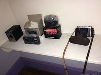Four vintage film cameras arranged on a white shelf, including the Ansco Shur Shot box camera, Photoflex MX-35 in box, Sears 35mm camera, and Kodak Retina folding camera with leather case