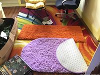 Displayed three rugs on hardwood floor and other room furniture. Large colorful area rug with sunburst design, orange and purple throw rugs.