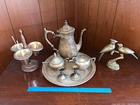 Photo of all items on wooden surface: silver plated tray, coffee pot, creamer, sugar bowl, egg cups, brass bird figurines, and brass candle holder with candles.