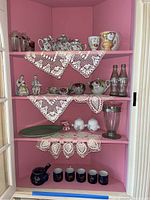 Full view of pink cabinet shelves with various tea sets, cups, figurines, glass vase, and CocaCola bottles arranged with crocheted doilies