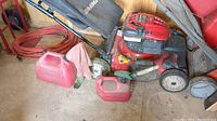 Lawn mower with two red plastic gas cans, orange extension cord in background.