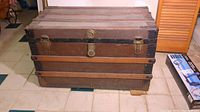 Front and side view of the large wooden trunk showing metal bands and leather handles. One handle is visibly broken.