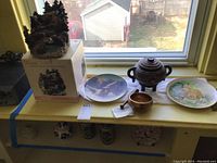 Battery operated water fountain atop its box, Avon Christmas plate, Staffordshire fox plate, lidded ginger jar vase on a windowsill