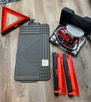 Photo showing two gray WeatherTech floor mats with grooved design, black carrying case open containing jumper cables and tools, and three orange triangular reflective emergency warning devices.