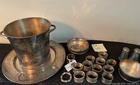 Photo showing silver ice bucket placed on a large silver charger with multiple silver napkin rings and a small bowl arranged in front, along with a pair of salt and pepper shakers.