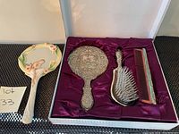 Silver plated 3 piece dresser set including ornate hand mirror, hairbrush with white bristles, and reddish comb inside purple satin-lined white box plus an additional ceramic decorative mirror.