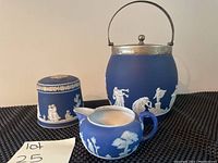 Front angled photo showing three pieces: large covered sugar bowl with silver trim and handle, small covered sugar bowl, and blue creamer. Highlights white classical reliefs on blue jasperware.