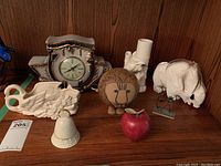 Overview of all items on wooden shelf including bell, clock, vase, swan planter, ivory figurines, cork lion, red apple and metal card holder
