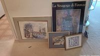 Five framed art pieces arranged leaning against wall and each other on carpet, showing various sizes and themes including Paris river scene and Italian synagogue poster