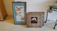 Two framed art pieces standing against wall, showing entire pieces.