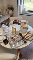 White ceramic casserole dish with lid, ceramic canisters with wooden lids, white ceramic custard cups stacked, metal muffin tins, brown ceramic pot, plates, cutlery set, kitchen scale, and glass coasters on round table by window.