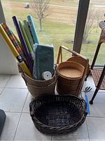 Full display of woven baskets and wrapped items including multiple rolls of colorful wrapping paper and green tissue paper inside a woven basket. The three-tier bamboo basket is shown with its handle upright, and a dark oval woven basket is in front.