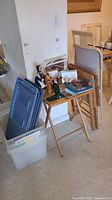 Photo showing two plastic storage bins with blue lids and three wooden TV trays leaning against a wall next to a wooden folding chair.