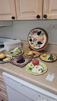 Overview of 7 ceramic fruit dishes and the ceramic pie plate with lid, all with colorful fruit motifs showing some sticky residue needing cleaning.