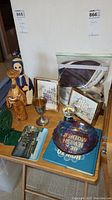 Overview of lot contents on wooden table, showing all items: figurines, bookend, goblet, framed art, glass bowl, book, and bag of yarmulkas.
