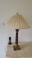Full view of one metal base lamp with cream pleated fabric shade and loop finial.