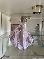Full view of porcelain figurine of girl wearing flowing pink skirt and holding bouquet, displayed in glass cabinet.