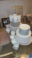 Full view of stacked white fine china including coffee cups, plates, bowls, creamer, and sugar bowl, arranged on a glass table with chairs in the background.