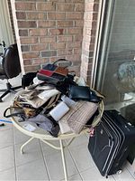 Photo showing a pile of multiple ladies purses and handbags on a round white metal outdoor table with a black rolling suitcase next to it.