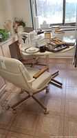 Photo showing vintage round wooden table with two matching beige leather chairs on wheels, set on tiled floor near window.