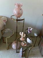 Overview of lot items displayed on glass and gold nested tables against wall: large pink ruffled glass vase on center table, small ceramic pitcher, cup and saucer, pink cactus candle, marble obelisk, round pink vase on floor next to tables, and glass bead display on tall stemmed glass vessel.