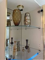 Shelf display showing large brown frosted vase with oval cutouts, tall metallic patterned vase, and several smaller vases in front with various patterns and styles.