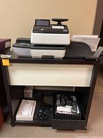 View of the white front panel and dark brown frame of the cabinet holding an office printer and containing electronic accessories in the lower shelf.